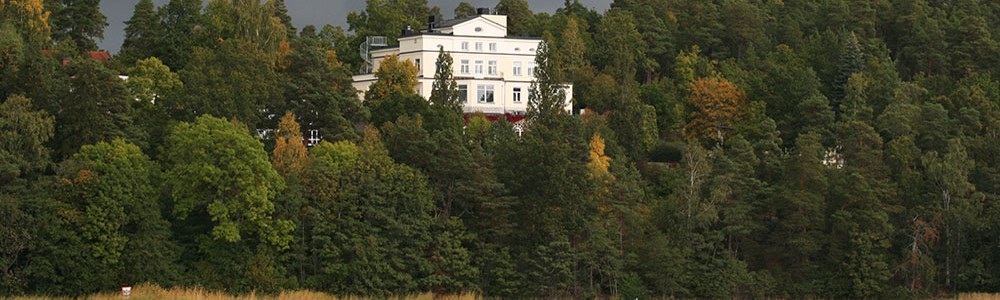 Vitt hus högst upp på en skogbeklädd sluttning  med strandkant nedtill