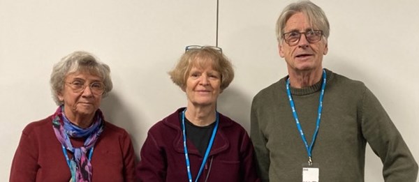 Två kvinnor och en man på rad mot en vit vägg.